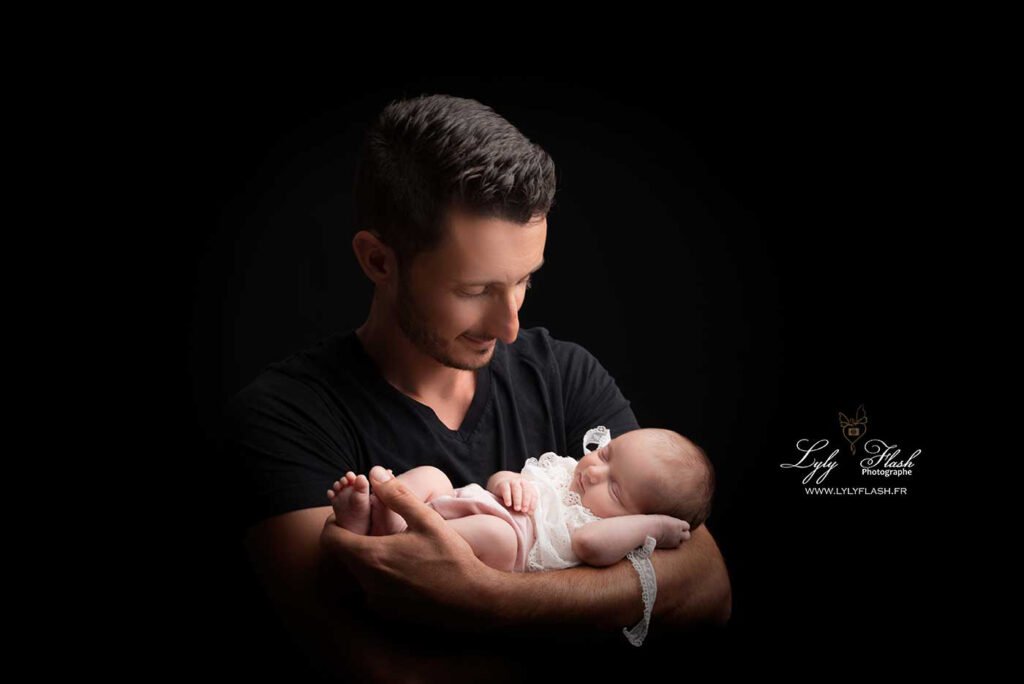 photo d une petite fille qui est née, il y a 5 jours dans les bras de son papa tout fière de la regarder et qui lui sourit en dormant, une photographie réalisée en studio photo par la photographe Lyly flash près de Correns