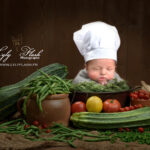photo de naissance d un bébé cuisinier, le bbé porte une grande toque blanche et dort entouré de tous les légumes provençale de saison
