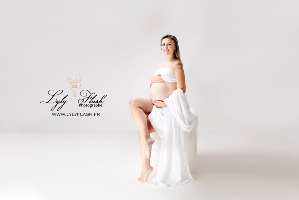 Roquebrune-sur-Argens – Dans une pose dynamique, la future maman est debout, ses bras levés au-dessus de sa tête, encadrant son visage. L’éclairage du studio met en valeur son ventre et ses traits doux, capturant la force et la grâce de la maternité.